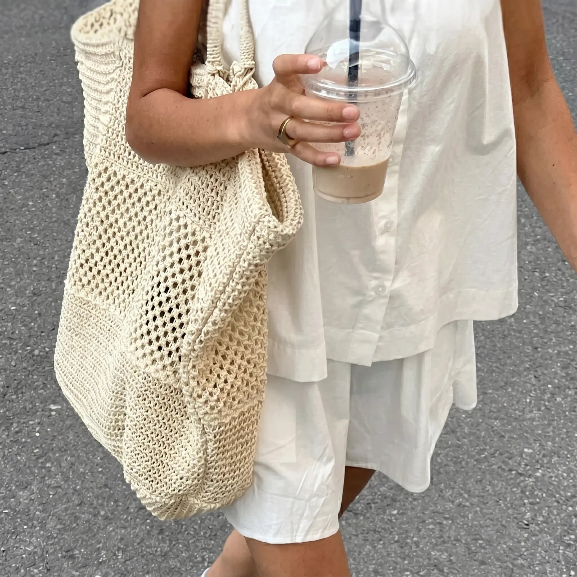 Woman wearing a crisp white 100% cotton camisole and shorts set, featuring a square neck cami with button details and spaghetti straps, styled casually outdoors with a large straw tote bag and a refreshing drink.