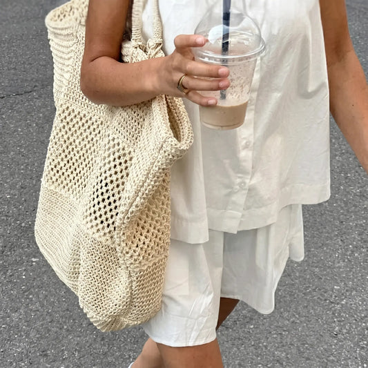 Woman wearing a crisp white 100% cotton camisole and shorts set, featuring a square neck cami with button details and spaghetti straps, styled casually outdoors with a large straw tote bag and a refreshing drink.