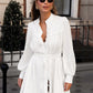 Woman wearing a white cotton linen mini dress with lantern sleeves, ruffled placket, and pom-pom hem, styled in an outdoor bohemian chic setting.