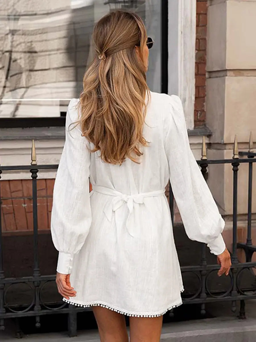 Back view of a white cotton linen mini dress featuring voluminous lantern sleeves, a cinched self-tie belt, and a delicate pom-pom trim along the hem.