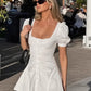 A white cotton mini dress featuring a square neckline, ruched bodice with pearlescent button front, and short puff sleeves, worn by a model outdoors in natural daylight.