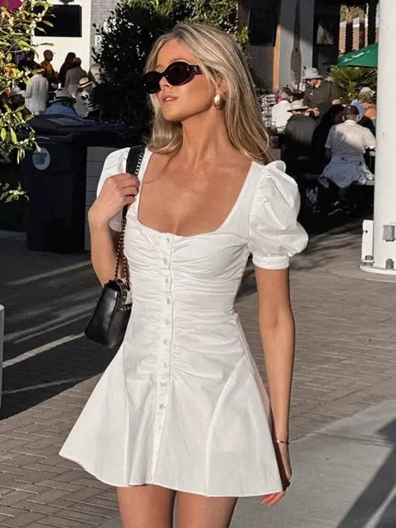 A white cotton mini dress featuring a square neckline, ruched bodice with pearlescent button front, and short puff sleeves, worn by a model outdoors in natural daylight.