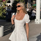 A white cotton mini dress featuring a square neckline, ruched bodice with pearlescent button front, and short puff sleeves, worn by a model outdoors in natural daylight.