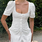 Elegant white cotton A-line mini dress with a square neckline, ruched bodice, and short puff sleeves, modeled outdoors.