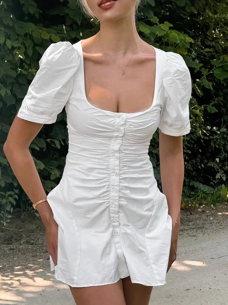 Elegant white cotton A-line mini dress with a square neckline, ruched bodice, and short puff sleeves, modeled outdoors.