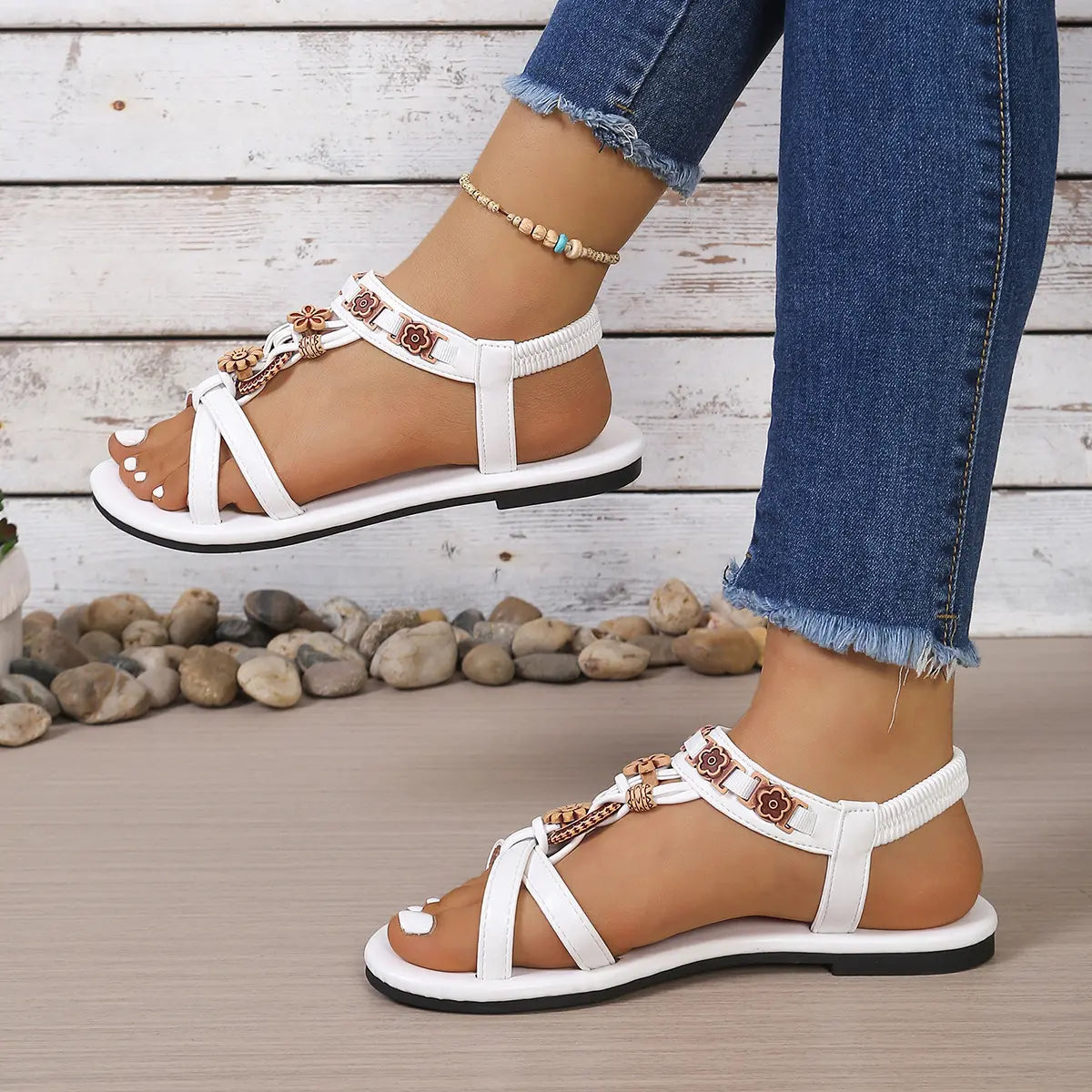 White embellished flat sandals with floral bead details and elasticated slingback straps, shown on a model wearing denim jeans in a lifestyle setting.