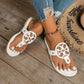 Woman's feet wearing white embellished flat thong sandals with ornate circular cut-out vamp and gold stud accents, styled with ripped jeans and an anklet, on a wooden floor beside a straw hat.