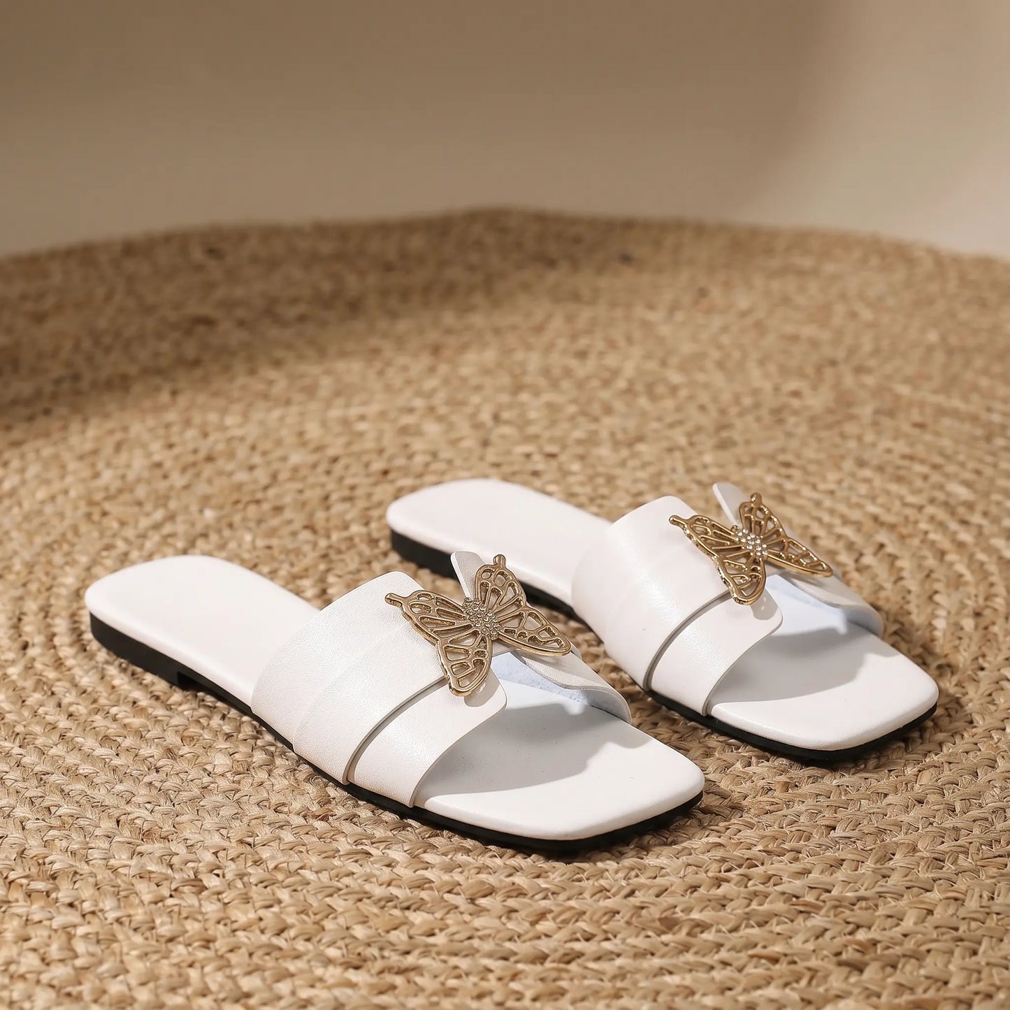 White flat slide sandals for women, featuring a square toe and a single wide strap adorned with an intricate gold-tone metal butterfly embellishment accented with crystals.
