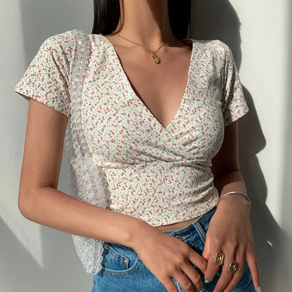 Model wearing a white Brivan floral faux-wrap V-neck crop top with short cap sleeves, styled with blue high-waisted jeans and gold jewelry.