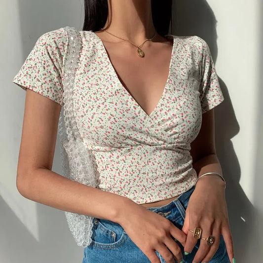Model wearing a white Brivan floral faux-wrap V-neck crop top with short cap sleeves, styled with blue high-waisted jeans and gold jewelry.