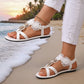 Elegant white floral flat sandals worn on a sandy beach at sunset, featuring delicate lace flowers, pearl accents, and a sparkling rhinestone oval buckle, paired with white jeans.