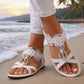 Elegant white floral flat sandals with pearl and rhinestone embellishments worn on a sandy beach with the ocean and sunset in the background.
