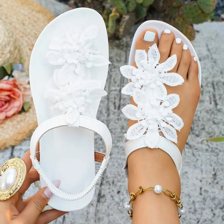 White flat thong sandals adorned with layered fabric floral appliqués, shown on a woman's foot with an elasticated slingback strap and stylish jewelry.