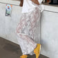 A woman models a flowing white floral lace maxi skirt paired with yellow sneakers, set against a textured concrete wall in a lifestyle setting.