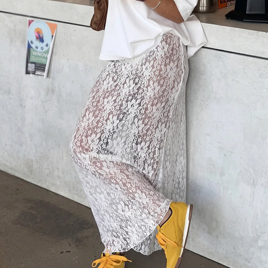 A woman models a flowing white floral lace maxi skirt paired with yellow sneakers, set against a textured concrete wall in a lifestyle setting.