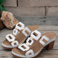 Pair of white heeled slide sandals with gold geometric hardware and white contrast stitching, displayed on a wooden surface. These platform sandals feature a chunky block heel and an open-toe design.