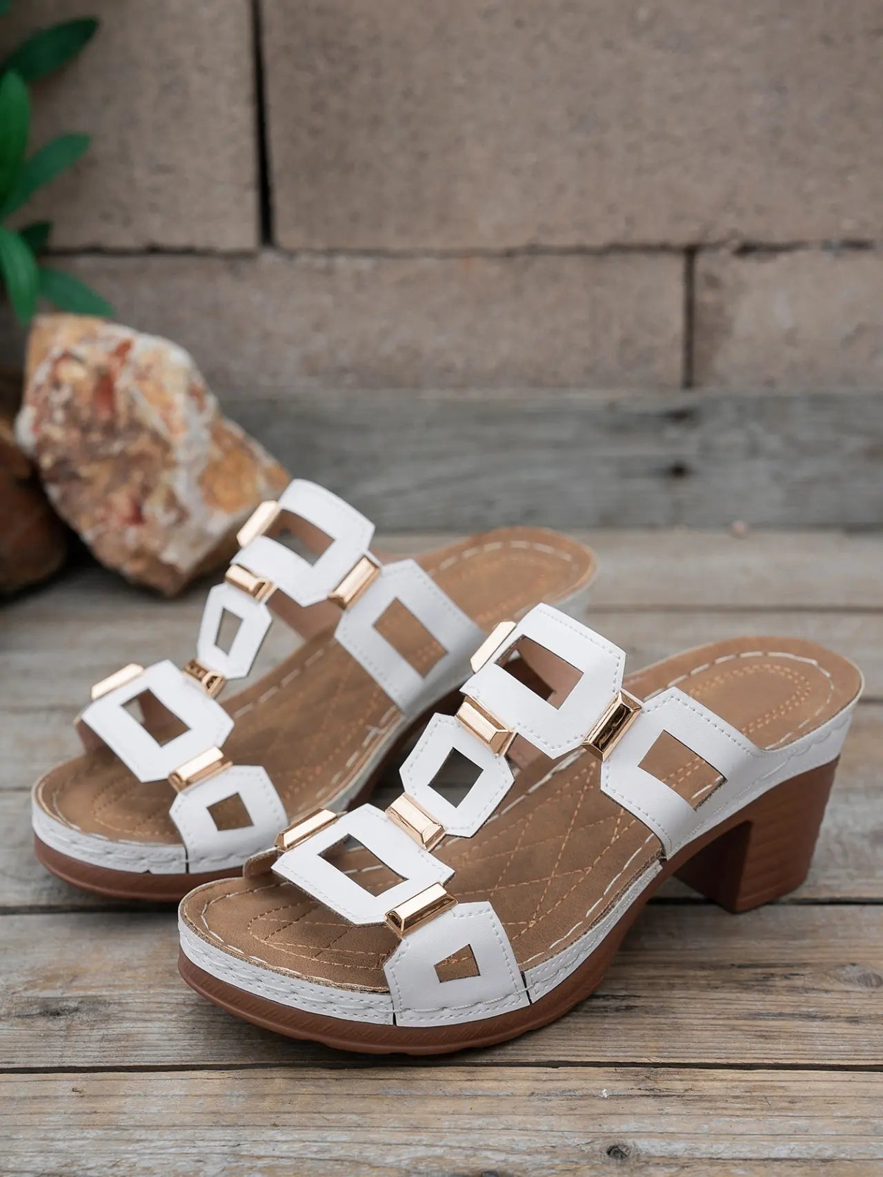 Pair of white heeled slide sandals with gold geometric hardware and white contrast stitching, displayed on a wooden surface. These platform sandals feature a chunky block heel and an open-toe design.