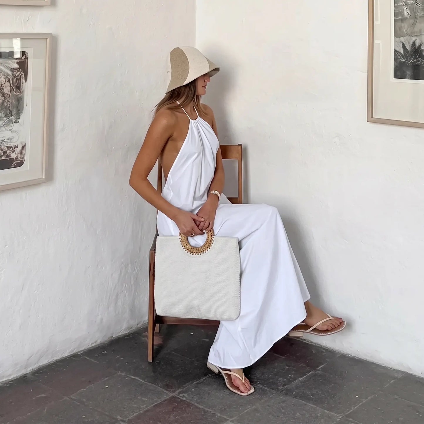 Woman wearing a flowing white bohemian halter maxi dress with an open back, seated with a woven bag and hat in a stylish resort setting.