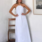 A smiling woman models a flowing white halter maxi dress with an open back, styled with a straw bucket hat and sandals in a chic, minimalist setting.