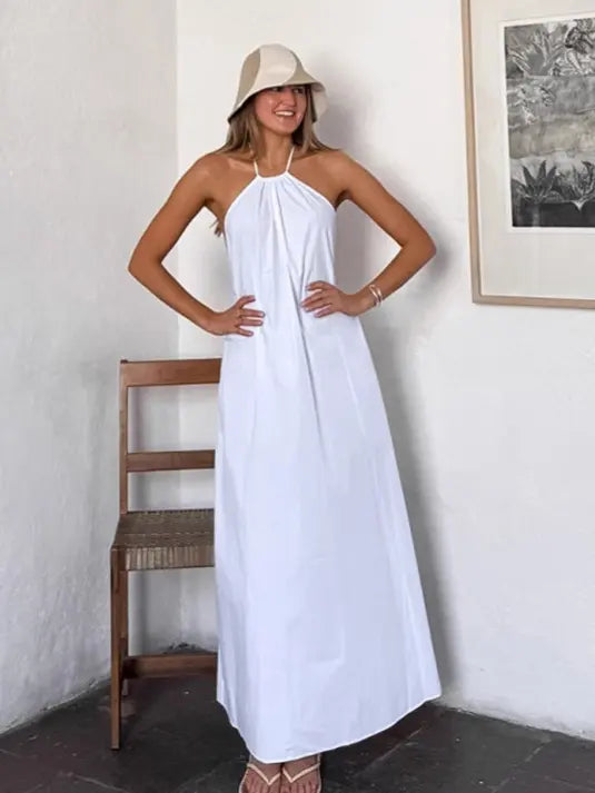 A smiling woman models a flowing white halter maxi dress with an open back, styled with a straw bucket hat and sandals in a chic, minimalist setting.
