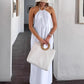 A woman wearing a flowing white halter maxi dress and a straw hat, holding a textured white tote bag, standing in a bright, minimalist hallway with white walls and dark tiled floors.