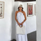 A woman models a flowing white halter maxi dress with an open back, styled with a straw hat and bag in a bright, art-filled resort interior.