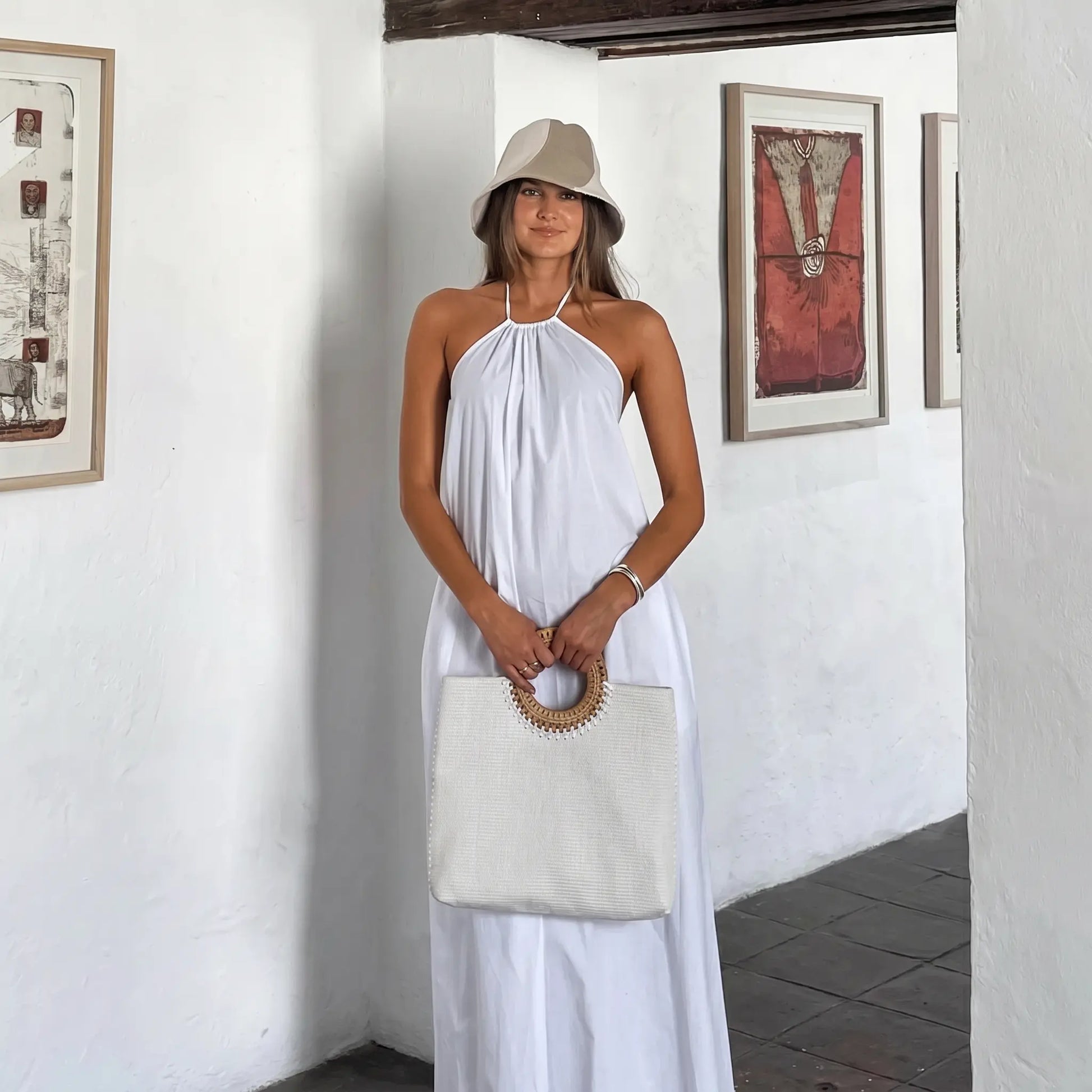 A woman models a flowing white halter maxi dress with an open back, styled with a straw hat and bag in a bright, art-filled resort interior.