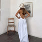 A woman stands with her back to the camera, wearing a flowing white halter maxi dress with an open back, styled in a minimalist room setting. white halter maxi dress open back bohemian resort wear.