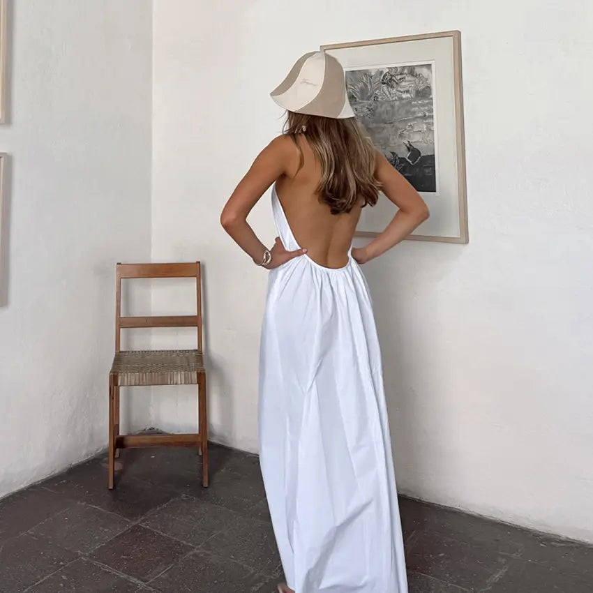 A woman stands with her back to the camera, wearing a flowing white halter maxi dress with an open back, styled in a minimalist room setting. white halter maxi dress open back bohemian resort wear.