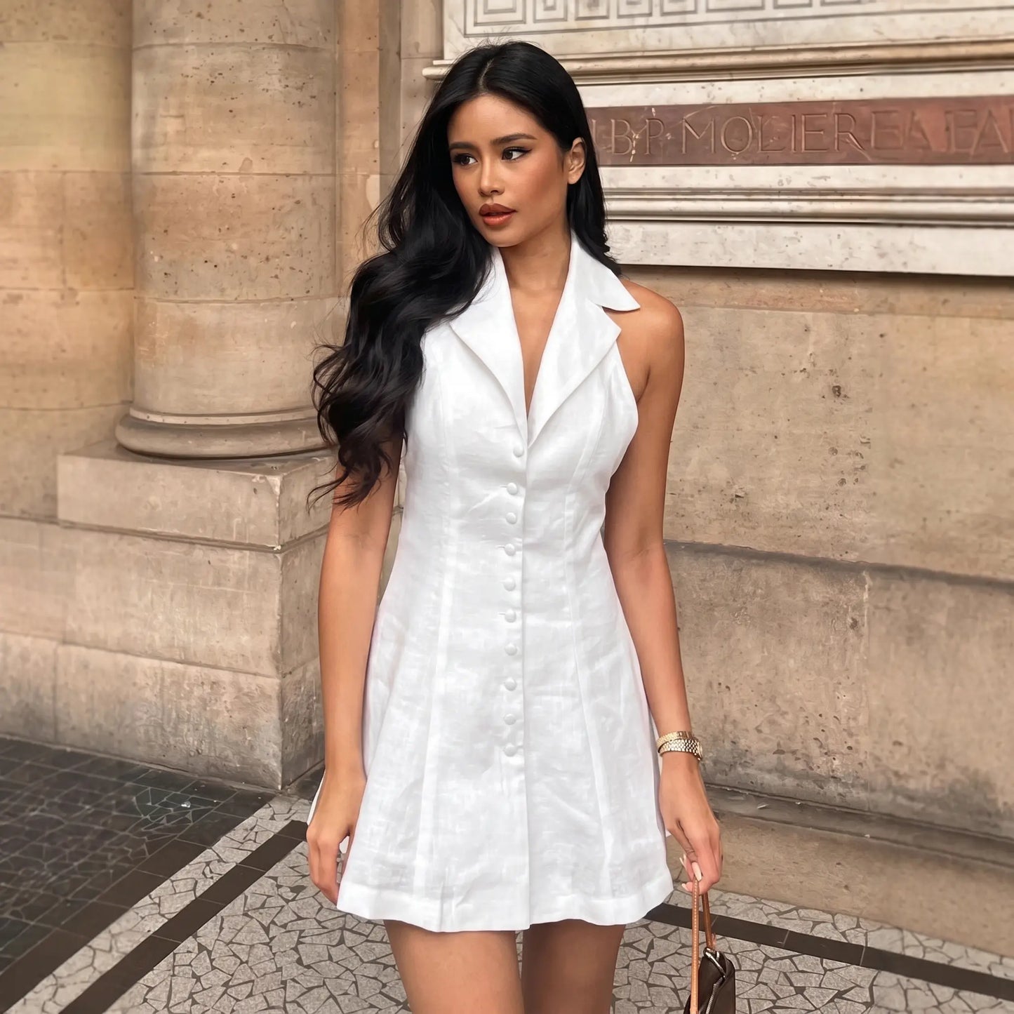 Woman wearing a white sleeveless linen-blend A-line mini dress with a halter neckline and button-front detail, standing outdoors against a stone building.