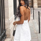 A model wears a white linen-blend A-line mini dress with a distinctive halter neckline and open back, posing outdoors against a classic stone building.