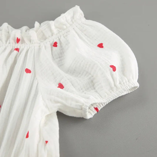 Close-up view of a white cotton gauze dress with an all-over red heart print, showcasing the ruffled off-shoulder neckline and puff sleeve detail.