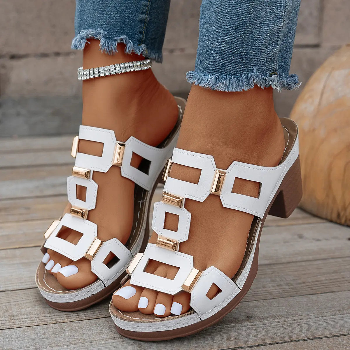 Women's white heeled slide sandals with geometric gold-tone hardware and a chunky block heel, styled with distressed denim and an anklet on a wooden deck.