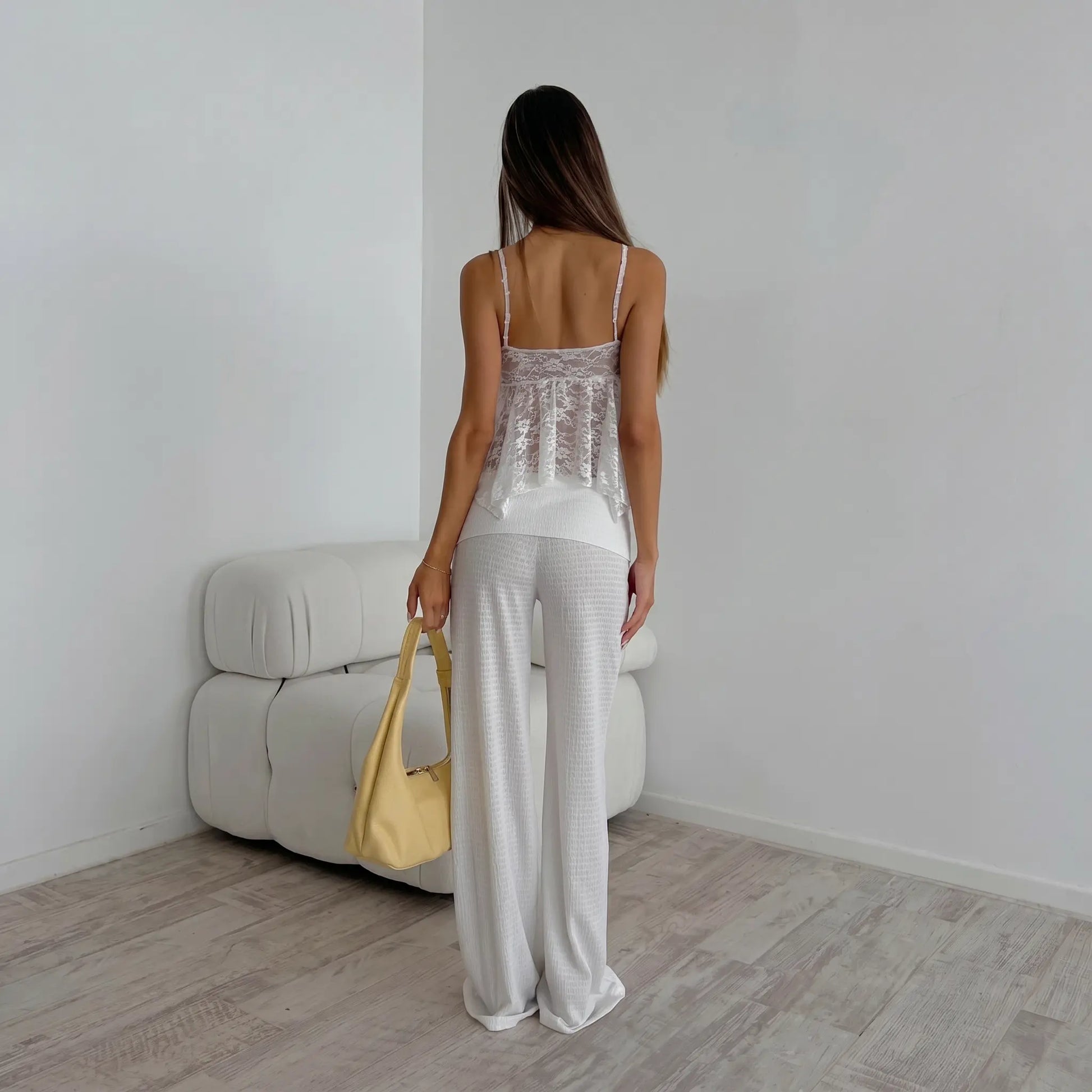 Woman wearing a white lace camisole and matching wide-leg pants, holding a yellow handbag, shown from the back in a minimalist studio setting.