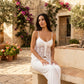A woman with dark hair sits in a sun-drenched courtyard wearing a white floral lace camisole with adjustable spaghetti straps and a tie-front detail, paired with white trousers.