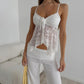 Woman wearing a white lace camisole with a tie-front detail and scalloped hem, paired with textured white pants. This elegant summer top showcases thin spaghetti straps and a romantic V-neckline.