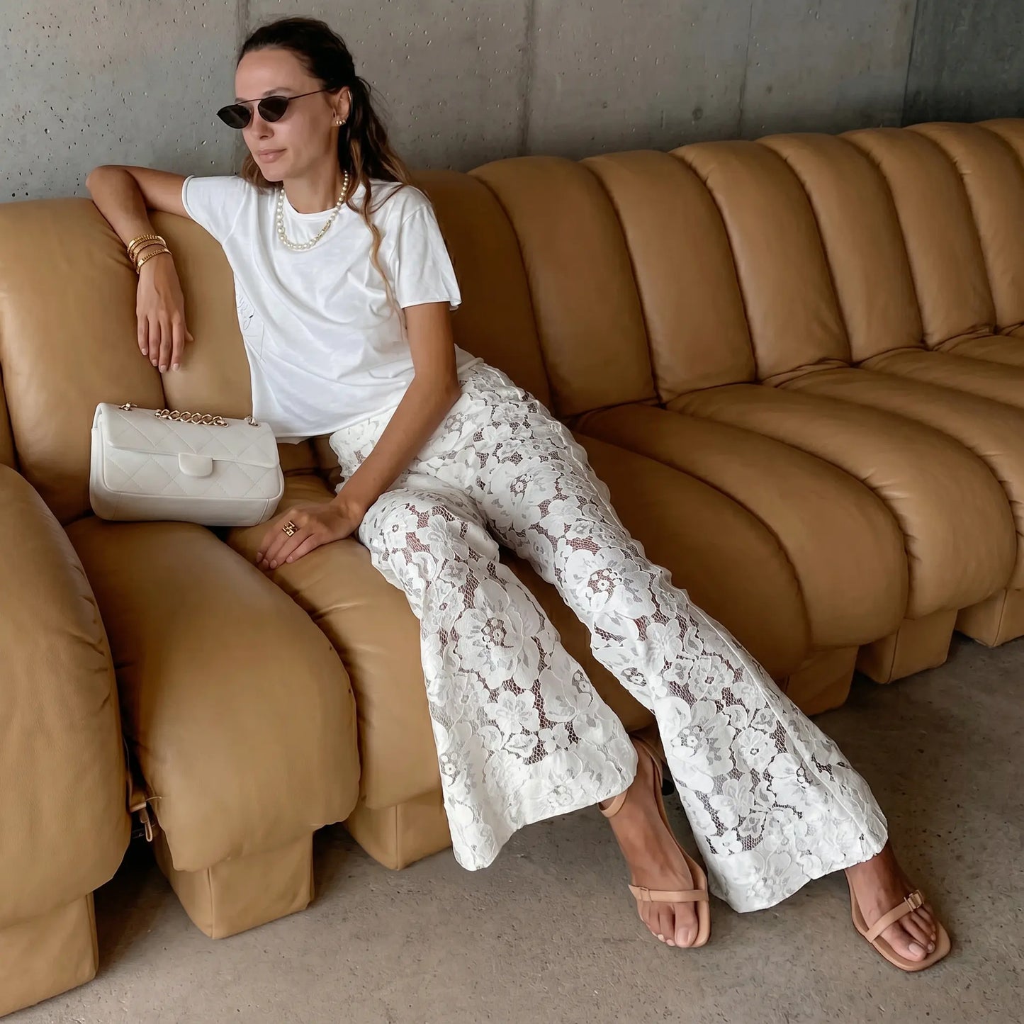 Model wearing elegant white lace flared trousers, showcasing their high-waist design, sheer bohemian pattern, and dramatic wide leg while seated on a sofa.