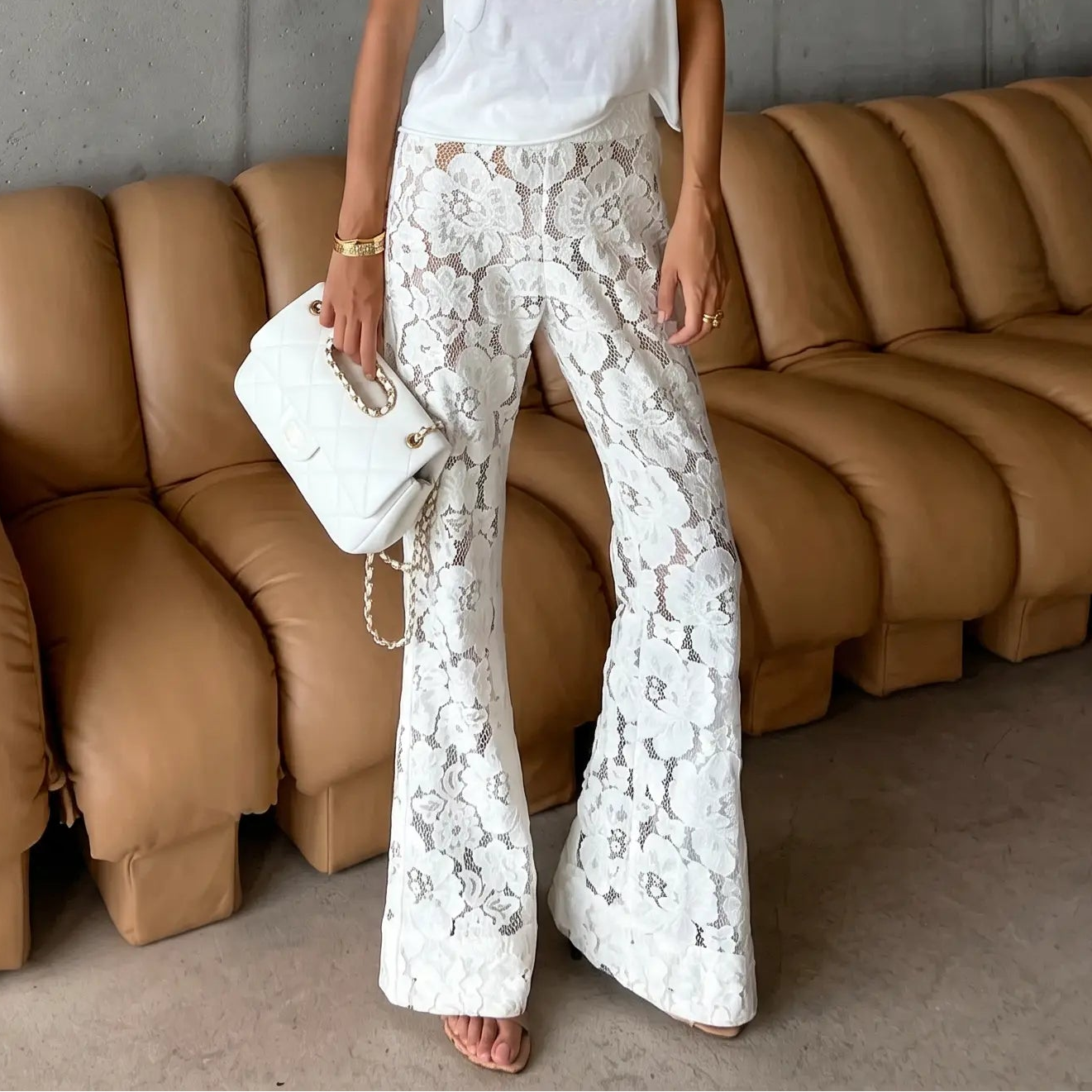 Model wearing white lace flared trousers and a white t-shirt, styled for a bohemian resort look with sunglasses and a handbag, posing against a concrete wall and leather sofa.