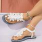 A pair of white flat thong sandals adorned with layered white lace floral embellishments and a white ankle strap with coiled elastic, shown on a model's feet with beach-themed anklets.