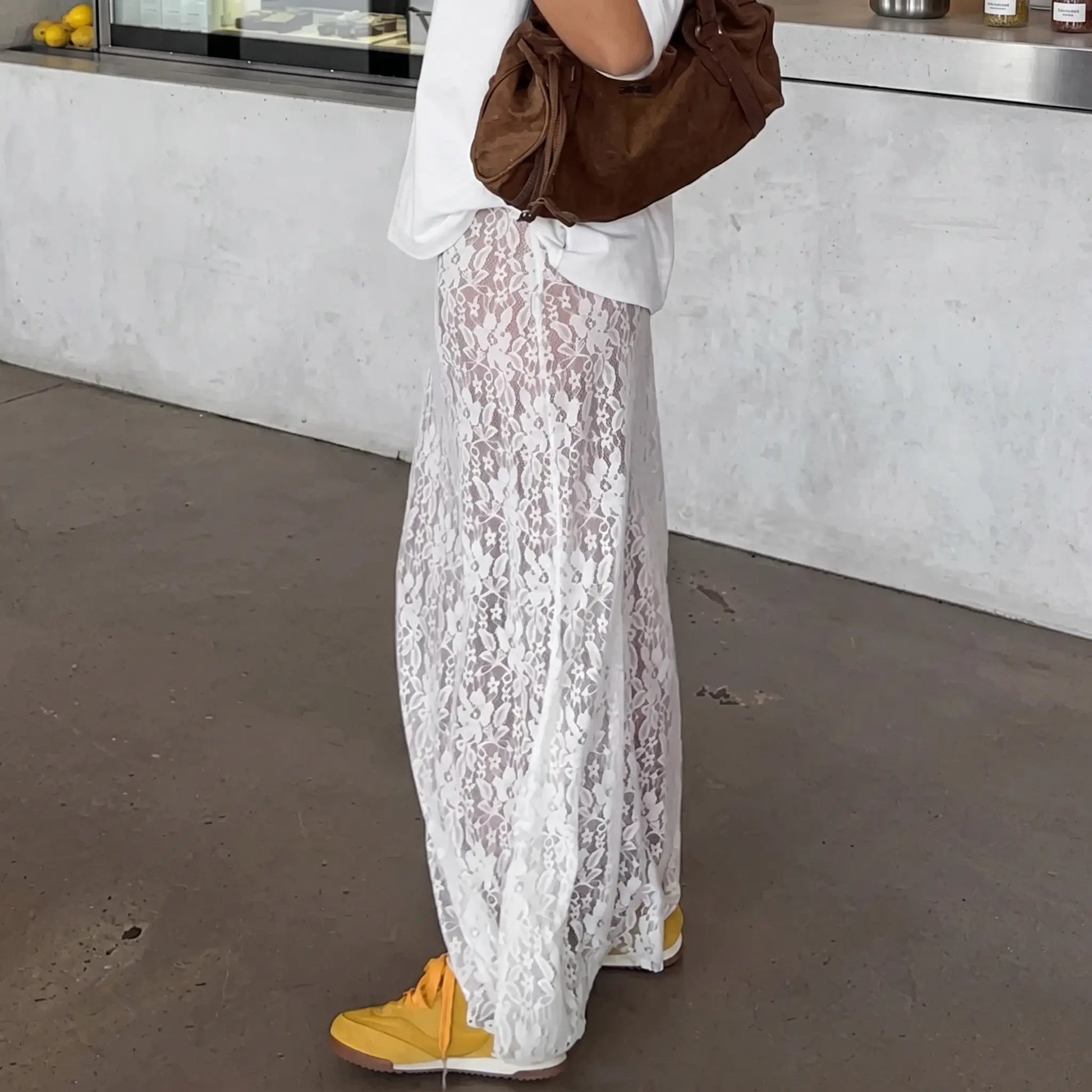 White floor-length A-line lace maxi skirt styled with a white t-shirt, brown bag, and yellow sneakers. The sheer lace allows visibility of the legs.