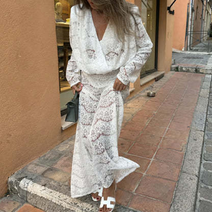 A woman models a romantic white floral lace two-piece set featuring a draped cowl neck top and flowing wide-leg trousers, styled with white sandals in an outdoor street setting.