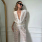 A woman models a white floral lace two-piece set featuring a draped cowl neck top with voluminous puff sleeves and high-waisted wide-leg pants, styled with sunglasses and a gold chain belt.