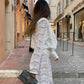 A woman walks down a cobblestone street wearing a flowing white floral lace two-piece set, featuring a draped cowl neck top with voluminous sleeves and wide-leg trousers. She carries a grey handbag.