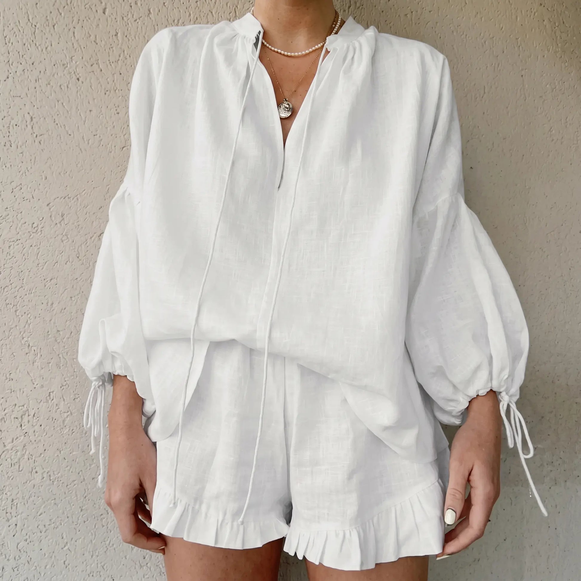 White linen-blend two-piece set featuring a loose V-neck blouse with voluminous lantern sleeves and ruffled hem shorts worn by a woman, showcasing elegant bohemian summer style.