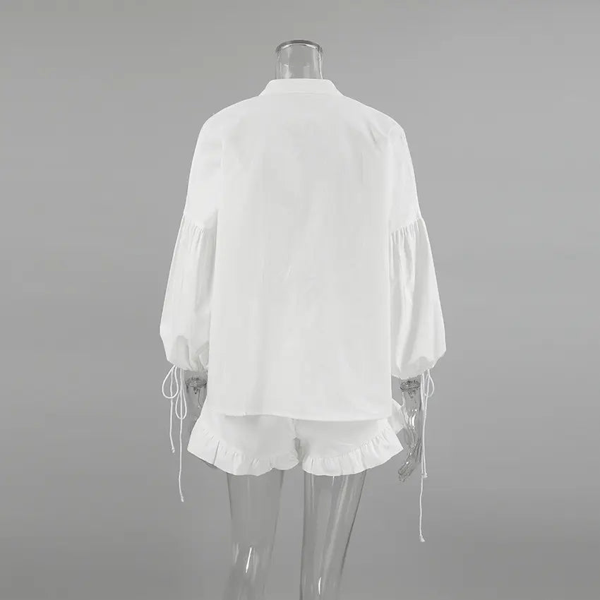 Back view of a white linen-blend set featuring a loose blouse with voluminous lantern sleeves and adjustable tie cuffs, paired with shorts that have a delicate ruffled hem. This ensemble showcases bohemian chic.