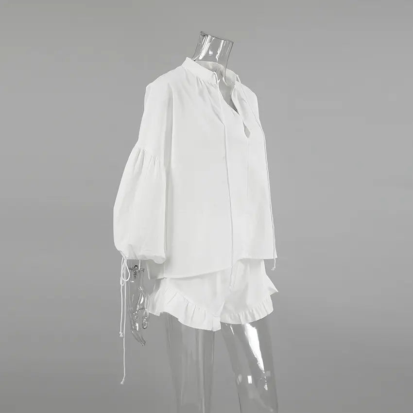 White linen blend two-piece set featuring a V-neck blouse with voluminous lantern sleeves and adjustable tie cuffs, paired with high-waisted shorts with a ruffled hem, shown from the side.