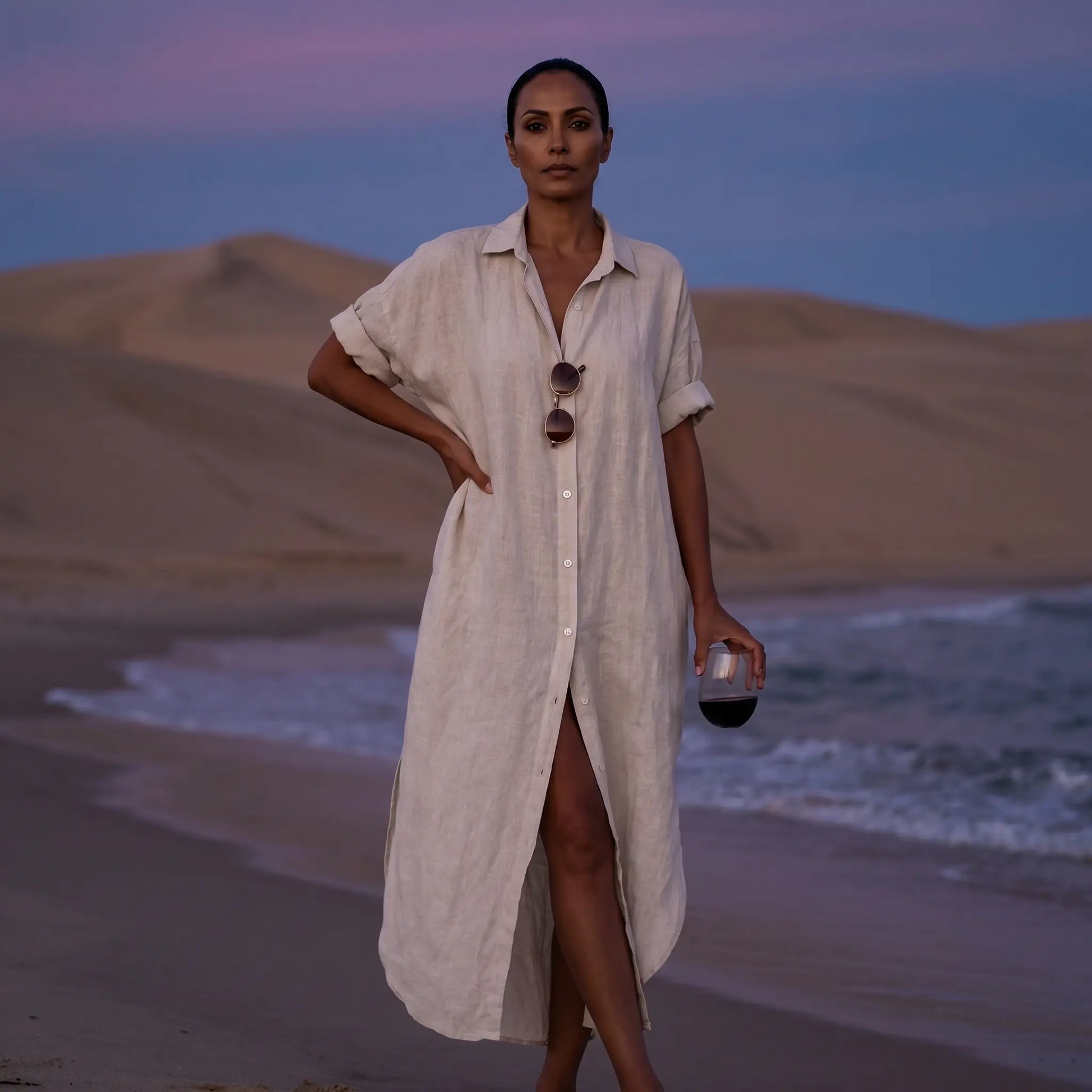 Model wears a pristine white linen blend maxi shirt dress with rolled sleeves and a collared neckline, posing on a twilight beach with ocean waves and sand dunes.