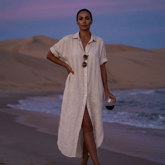 Model wears a pristine white linen blend maxi shirt dress with rolled sleeves and a collared neckline, posing on a twilight beach with ocean waves and sand dunes.