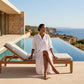 Woman in a white linen-blend maxi shirt dress with rolled sleeves, sitting elegantly on a poolside lounge chair overlooking the ocean on a bright day.