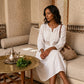 Model wearing a Kivren Pure White Bohemian Embroidered Linen Midi Dress, sitting on a light-colored sofa in a traditional, rustic indoor setting. The dress features a notched V-neck, 3/4 sleeves, and an elegant geometric embroidered empire waist. A brass table with a teapot and mint tea is in the foreground, evoking a serene moment.